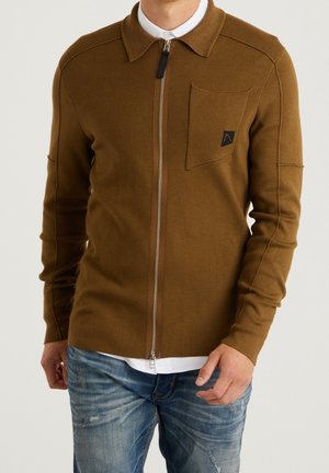 Mężczyzna w brązowym swetrze na zamek z kieszenią na piersi, noszący biały t-shirt i niebieskie sprane dżinsy, stojący na jednolitym tle.