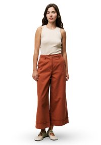 Débardeur beige à côtes, pantalon large rouille avec ourlet à franges et chaussures beiges à lacets. Le mannequin se tient contre un fond blanc.