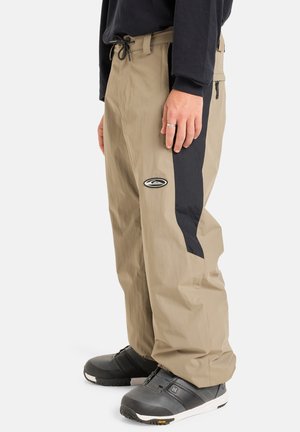Pantalons imperméables beiges avec un panneau latéral noir, taille élastique avec cordon de serrage et un petit logo sur la cuisse. Matériau durable et coupe ample.