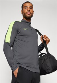 Donkergrijze lange mouw sporttop met een hoge kraag en contrasterende neon groene schouderaccenten, met een Nike-logo op de borst.