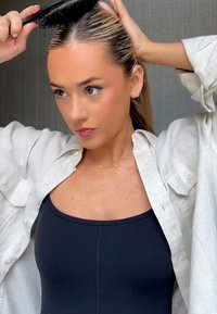 Schwarzer Kamm in einer Hand, glattes blondes Haar nach hinten frisiert, trägt ein schwarzes Tanktop und ein lockeres cremefarbenes Hemd. Neutraler Hintergrund.