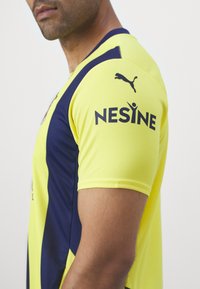 Gelbes und marineblaues Kurzarmtrikot mit dem "Nesine"-Logo und Puma-Branding, aus atmungsaktivem Material mit strukturierter Oberfläche.