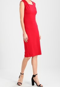 Robe rouge longueur genou avec un décolleté carré et des manches courtes. Confectionnée en tissu lisse, elle présente une taille cintrée et une silhouette droite.