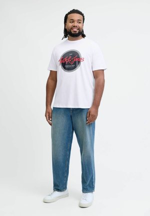Witte T-shirt met zwart cirkelvormig grafisch ontwerp en rode tekst. Gepaarde met losse blauwe jeans en witte sneakers. Eenvoudig, casual ontwerp.