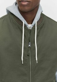 Grön bomberjacka med dragkedja, featuring en grå hoodie under. Dragsko i vitt, med en ribbad krage och textur.