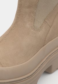 Botín de ante beige con paneles laterales de malla y una suela de goma gruesa en color beige. Presenta detalles cosidos y un diseño de punta redondeada.