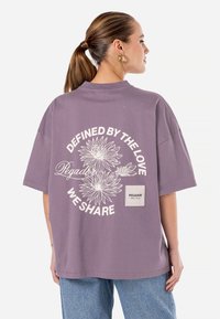 Purpuriu per didelis T-shirt, pagamintas iš medvilnės, su gėlių grafika ant nugaros ir logotipo užsiuvimu šone. Trumpomis rankovėmis ir apvalia iškirpte.