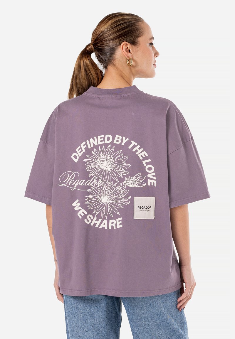 Purpuriu per didelis T-shirt, pagamintas iš medvilnės, su gėlių grafika ant nugaros ir logotipo užsiuvimu šone. Trumpomis rankovėmis ir apvalia iškirpte.