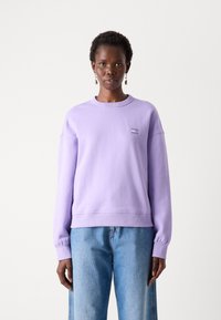 Felpa di cotone lavanda con collo rotondo, polsini e orlo a coste. Presenta un piccolo dettaglio grafico sul petto. Abbinata a jeans azzurri chiari.