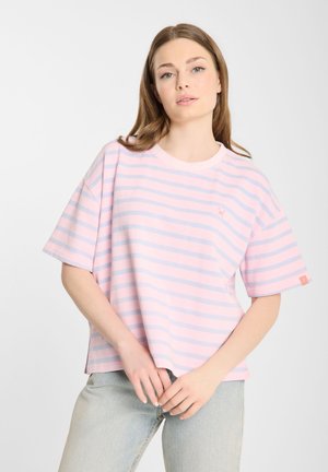 Jonge vrouw draagt een oversized roze en lichtblauw gestreept T-shirt en lichtgewassen jeans, staand tegen een witte achtergrond.