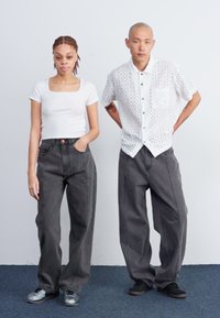 Due persone stanno fianco a fianco; una indossa una maglietta bianca aderente e jeans grigi larghi, l'altra una camicia bianca perforata e pantaloni grigi larghi.