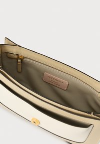 Interieur van een handtas in roomkleurig en beige leer met gouden beslag, inclusief een ritssluitingvak en een label met de tekst "Coccinelle Authentiek Italiaans Leer."