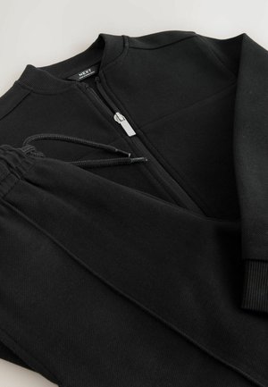 Schwarzer Sportanzug aus strukturiertem Stoff. Enthält eine Jacke mit Reißverschluss und einem gerippten Kragen sowie eine Hose mit Kordelzug und elastischen Bündchen.