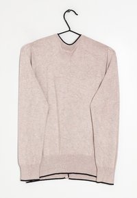 Ljus beige långärmad tröja med svart kant på kragen och i fållen hängande på en svart klädhängare mot en vit bakgrund.