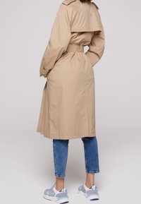 Cappotto trench beige con vita aderente, maniche lunghe e spacco posteriore. Indossato su jeans blu e sneakers chiare. Texture liscia, design classico.