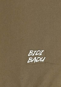 La tela de color verde oliva presenta una superficie texturizada con un logotipo blanco de "BIDI BADU" exhibido de manera destacada en la esquina inferior derecha.