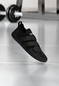 Schwarzer Sportschuh mit einem dehnbaren Obermaterial, doppelten Klettverschlussriemen, strukturiertem Sohlenprofil und minimalistischen Design. Besitzt dezente Branding-Elemente.