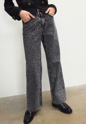 Person trägt hoch taillierte, weit geschnittene graue Jeans mit Leopardenmuster, hat die Hände in den Taschen, kombiniert mit schwarzen Loafers und einer kurz geschnittenen schwarzen Jacke.