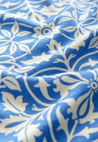 Blauer Stoff mit einem weißen Blumenmuster, das sich aus ineinander verschlungenen Blättern und Blumen zusammensetzt. Weiche Textur mit leicht zerknittertem Aussehen.