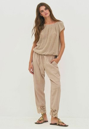 Vrouw met lang haar, gekleed in een losse beige top, lichtbruine broek met trekkoord en kanten boorden, en gespen sandalen, staand met één hand in de zak.