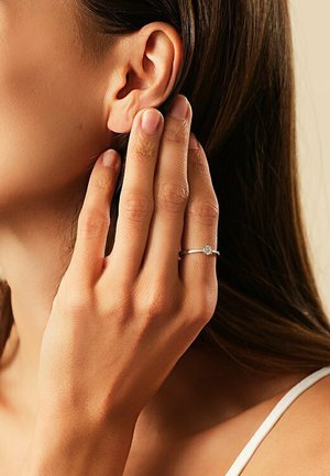 Silberring mit einem kleinen, klaren Stein, auf einem schlanken Band gefasst. Am Ringfinger getragen, die Hand in der Nähe des Ohrs positioniert, wodurch der natürliche Hautton zur Geltung kommt.