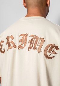 Cremefarvet kortærmet t-shirt med stort, udsmykket brunt broderi, der staver "CRIME" på ryggen. 100% bomuldsstof med en glat overflade.