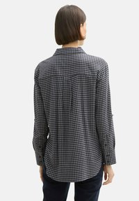 Chemise �à carreaux noir et blanc avec des manches longues, des poignets à boutons et un ourlet légèrement arrondi. La vue de dos montre un détail de pattes d'épaule et une texture de tissu douce.
