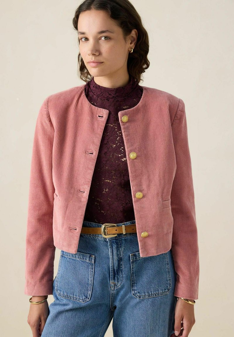 Veste en velours côtelé rose avec un col rond, dotée de boutons dorés et de manches longues, portée sur un haut en dentelle et un jean taille haute bleu.