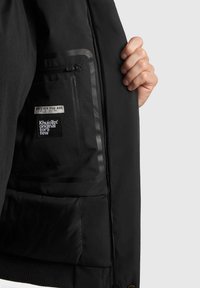 Juodas striukės pamušalas su užtrauktuko kišenę, turinčia skaidrų atvartą ir prekės ženklų etiketėmis. Medžiaga yra lygi su matinio finišo.