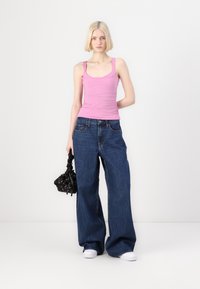 Top de tirantes ajustado rosa, pantalones vaqueros anchos de mezclilla azul, zapatillas blancas y un bolso negro con acentos florales. El fondo es blanco liso.