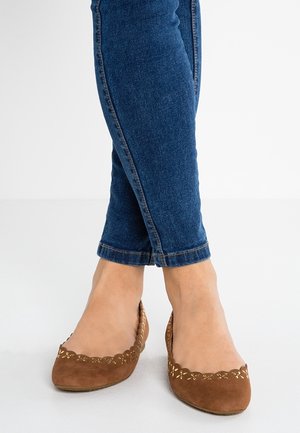 Pies con bailarinas de ante marrón con patrones decorativos recortados, combinados con vaqueros pitillo azul oscuro remangados sobre un fondo blanco.