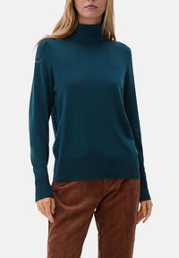 Pull à col roulé teal à manches longues, texture douce et coupe ajustée ; porté avec un pantalon en velours côtelé marron.