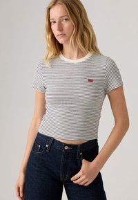 Striped short-sleeve T-shirt in white and navy, featuring a ribbed texture and a red logo label. Paired with dark blue high-waisted jeans.