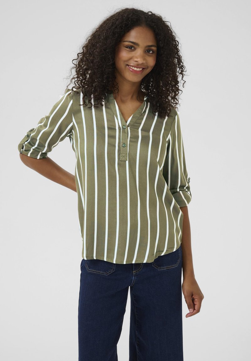 Femme souriante aux cheveux bouclés portant un chemisier à rayures verticales olive et blanches ainsi qu'un jean bleu foncé, posant avec une main sur la hanche.