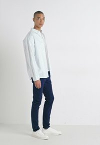 Chemise rayée à manches longues dans des couleurs pastel, associée à un pantalon bleu marine et des sneakers blanches, avec une coupe décontractée et un col boutonné.