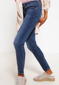 Osoba v úzkých modrých džínách, béžových semišových teniskách, světle růžovém svetru a béžovém kabátu, pózuje z profilu na bílém pozadí.