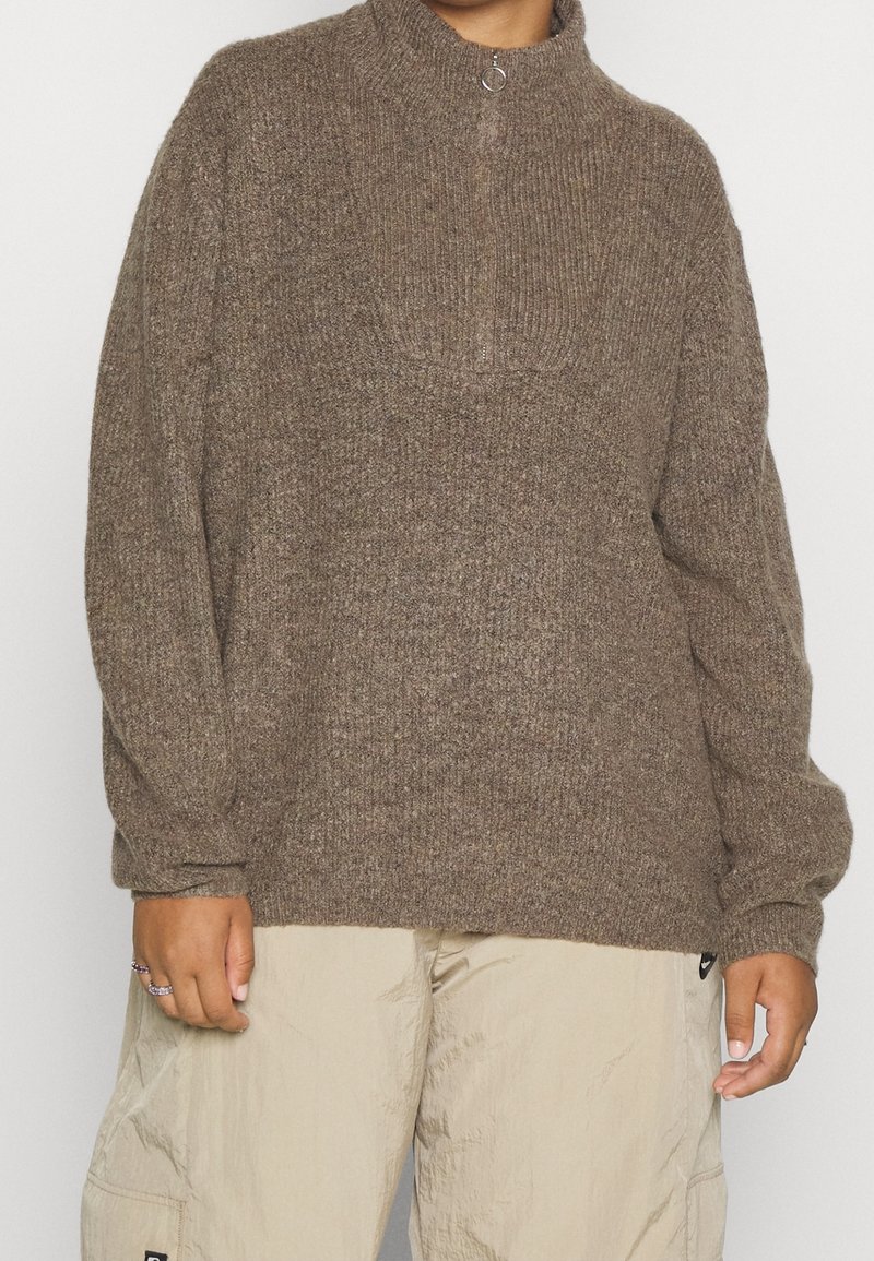 Pull marron tricoté avec un col montant et une fermeture demi-zip. Présente des manches surdimensionnées et un motif texturé. Associé à un pantalon cargo de couleur claire.