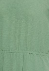 Tissu en mélange de coton vert, texturé avec un léger éclat, présente une taille élastiquée et un détail froncé à l'ourlet.