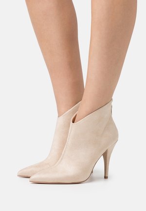 Bottines beige en daim à bout pointu avec talons hauts fins portées sur des jambes nues sur fond blanc.