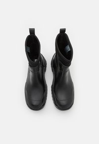 Une paire de bottes de pluie noires à la cheville avec des pattes de tirage, des semelles texturées et une finition mate lisse, vues d'en haut sur un fond blanc.