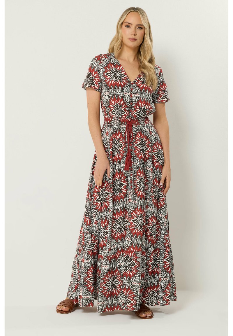 Femme debout portant une robe maxi à manches courtes avec un motif rouge, blanc et noir et une taille nouée, associée à des sandales marron.