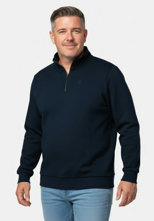 Mand med kort gråt hår iført en marineblå kvart-lynlås trøje og lyseblå jeans, stående mod en ensfarvet hvid baggrund.