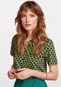 Blouse à manches courtes avec une base verte et un motif de feuilles noires. Le tissu présente une texture lisse avec un petit col.