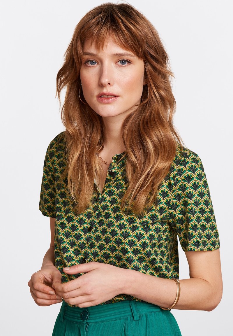 Blouse à manches courtes avec une base verte et un motif de feuilles noires. Le tissu présente une texture lisse avec un petit col.