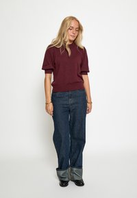 Burgundska kratka pletena majica s kratkimi rokavi v kombinaciji s temno modrimi širokimi hlačami. Hlače imajo zavihan rob, ki razkriva svetlejši denim. Črne čevlje.