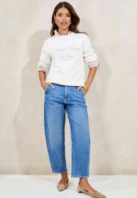 Felpa bianca con testo rosa, maniche a righe che sbucano. Jeans a gamba larga azzurri e ballerine beige completano l'outfit.