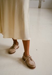 Zapatos de ante beige con cordones, de punta redonda y suela texturizada marrón, llevados con una falda de color crema y fluida, sobre un suelo liso y claro.