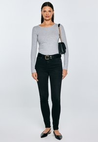 Sieviete ar taisniem tumšiem matiem, tērpusies pelēkā garroku blūzē, melnās augstās vidukļa plānas džinsās, melnā jostā, melnās sandālēs ar siksniņu un melnu plecu somu.