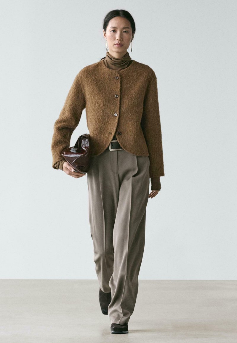 Brauner, strukturierter Cardigan mit Knöpfen, getragen über einem braunen Rollkragenpullover und beigen Hosen. Das Modell hält eine glänzende burgunderrote Tasche. Neutraler Hintergrund.