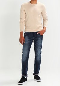 Beige Strickpullover, taillierte Passform, kombiniert mit dunkelblauen Denim-Jeans, leicht abgenutztem Finish und schwarzen Sneakers mit weißen Sohlen.